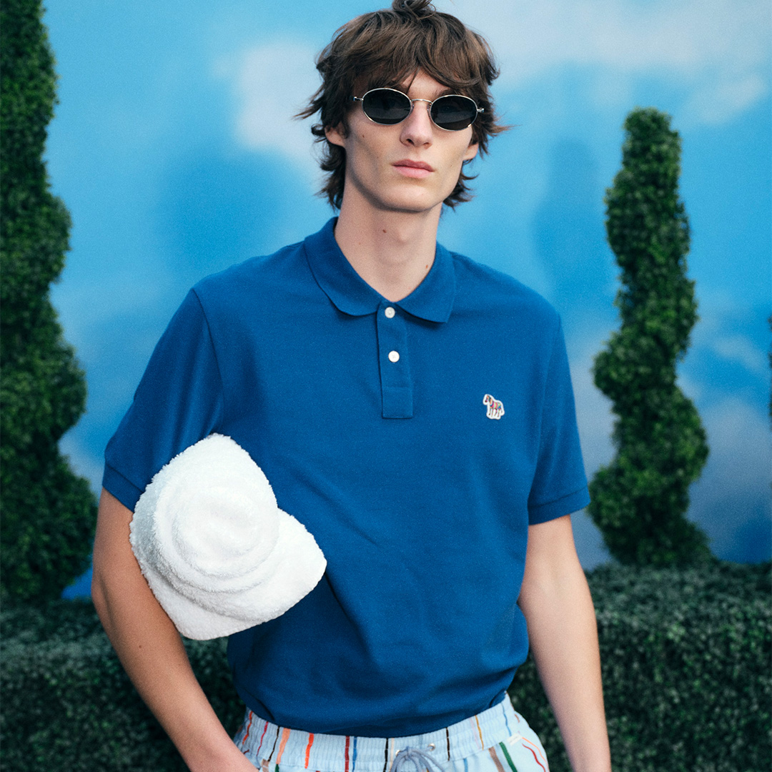 A person in a blue shirt and white shorts stands next to a white chair, set against a wooden backdrop.