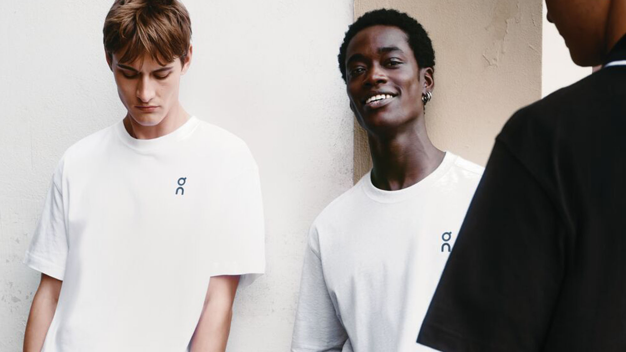 Three people in casual white and black T-shirts stand against a wall. Two are facing forward, one is turned away.