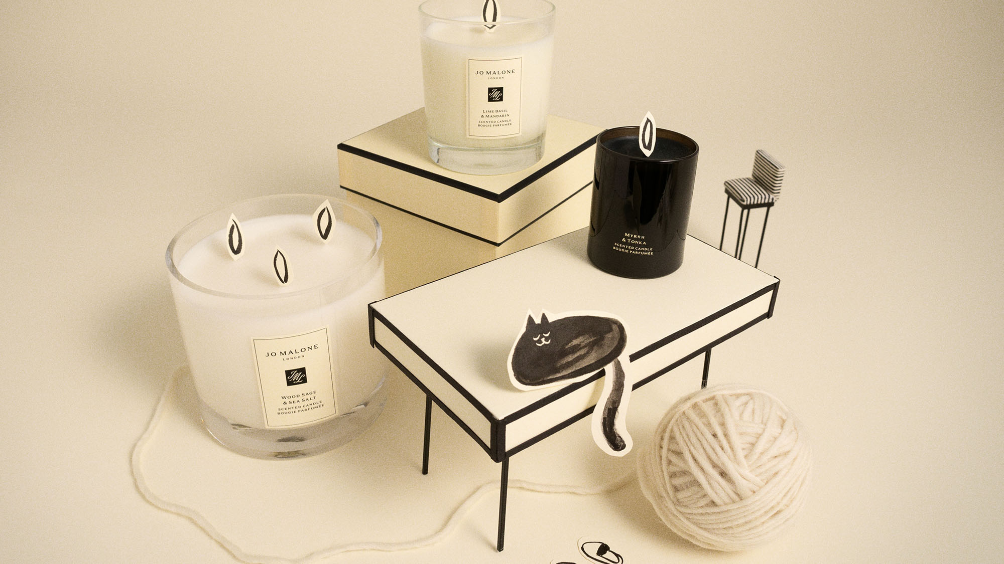 Jo Malone London home fragrance collection featuring multiple scented candles in glass containers, stacked books, black decorative cat, striped matches, and cream yarn on neutral background