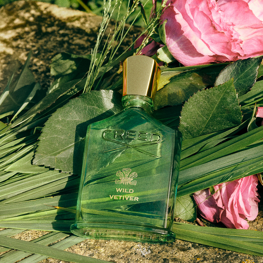 Creed Wild Vetiver fragrance bottle on sand with pink flowers and green foliage in sunlight