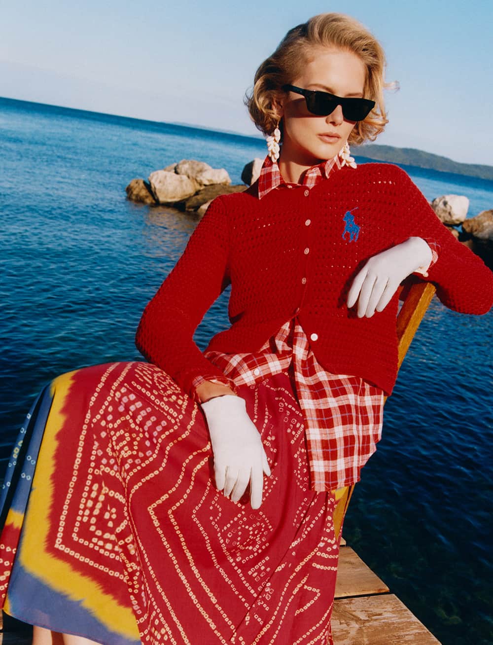 A person sits on a dock by the sea, wearing a red cardigan with a blue logo, a red and white checked shirt, a patterned maxi skirt, white gloves, and black sunglasses. The background features calm water and distant hills under a clear sky.