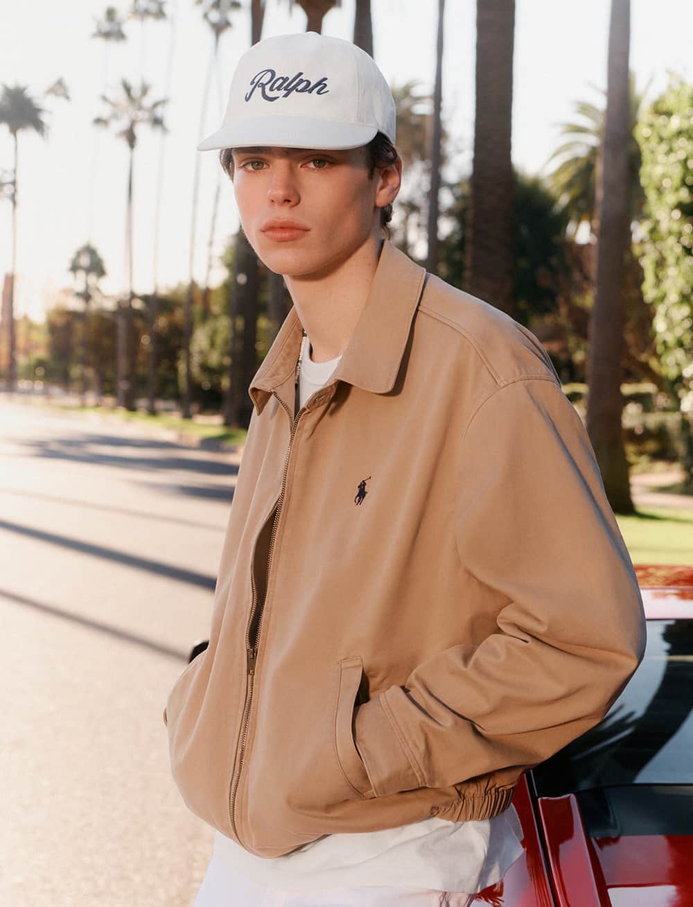 A person wearing a beige jacket and a white cap stands by the side of a street lined with tall palm trees. The sunlight casts long shadows, and a red car is partially visible in the foreground.