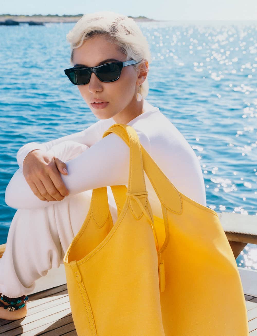 A person with short hair and sunglasses sits on a boat, dressed in a white outfit. They are holding a large yellow bag. The backdrop is a sparkling blue sea under a clear sky.