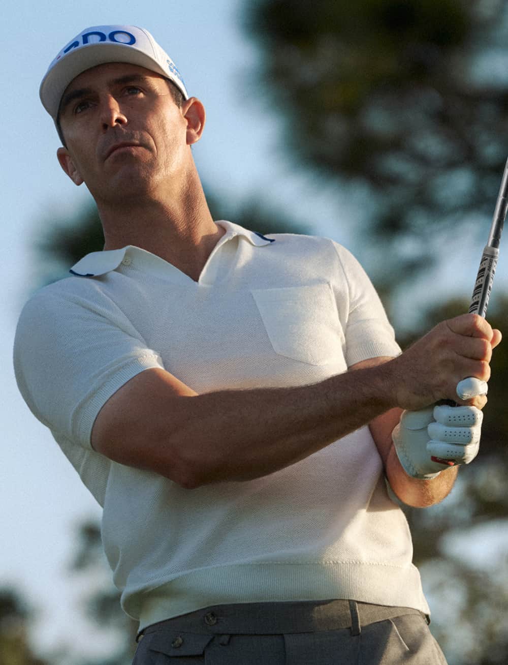 A golfer wearing a white shirt and cap focuses on his swing, holding a golf club. The background shows blurred greenery.