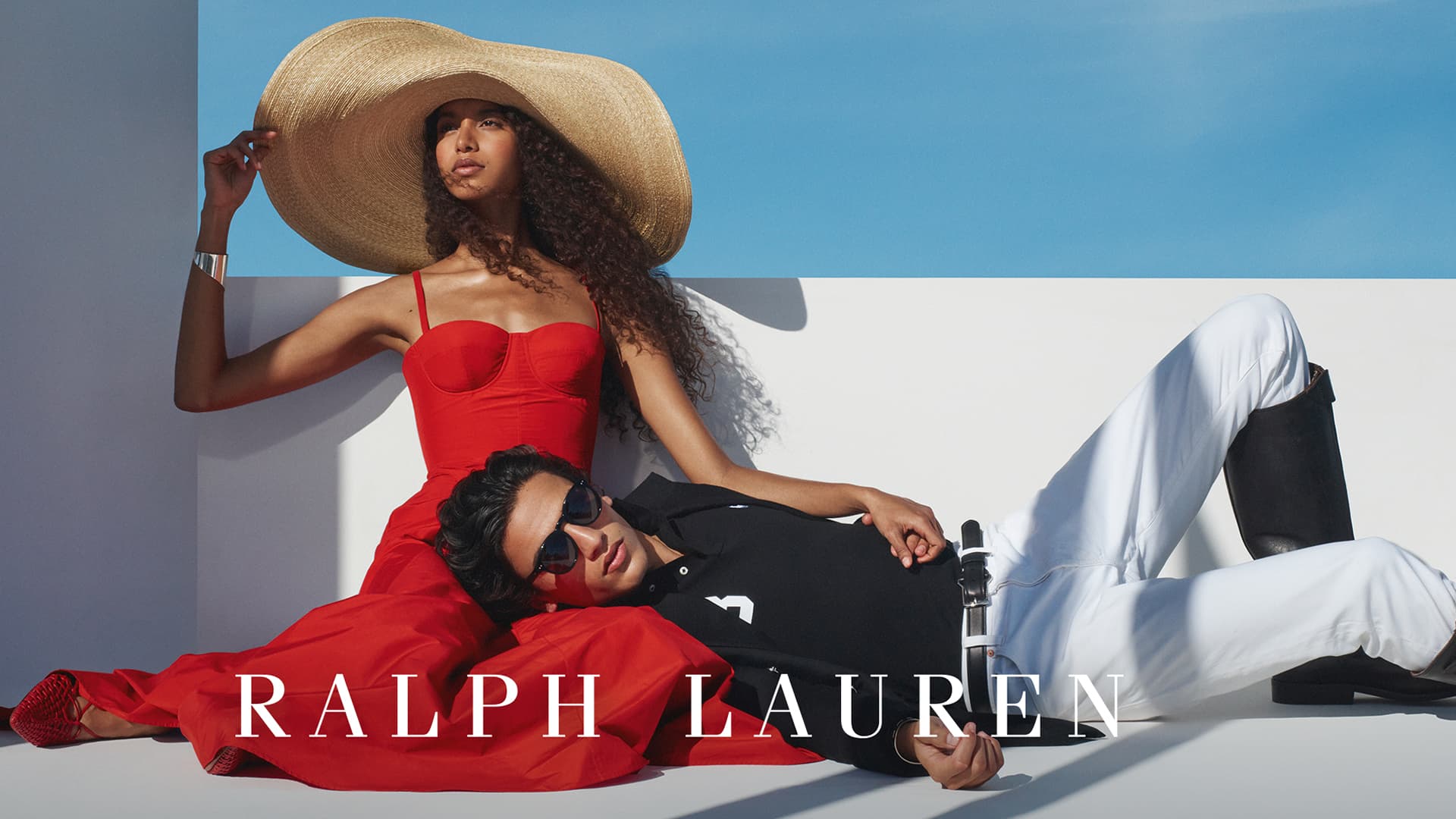 A woman in a red dress and wide-brimmed straw hat stands confidently against a white wall. Beside her, a man in white trousers and a black polo shirt reclines, resting his head on her lap. Both are wearing sunglasses. The Ralph Lauren logo is displayed across the image.