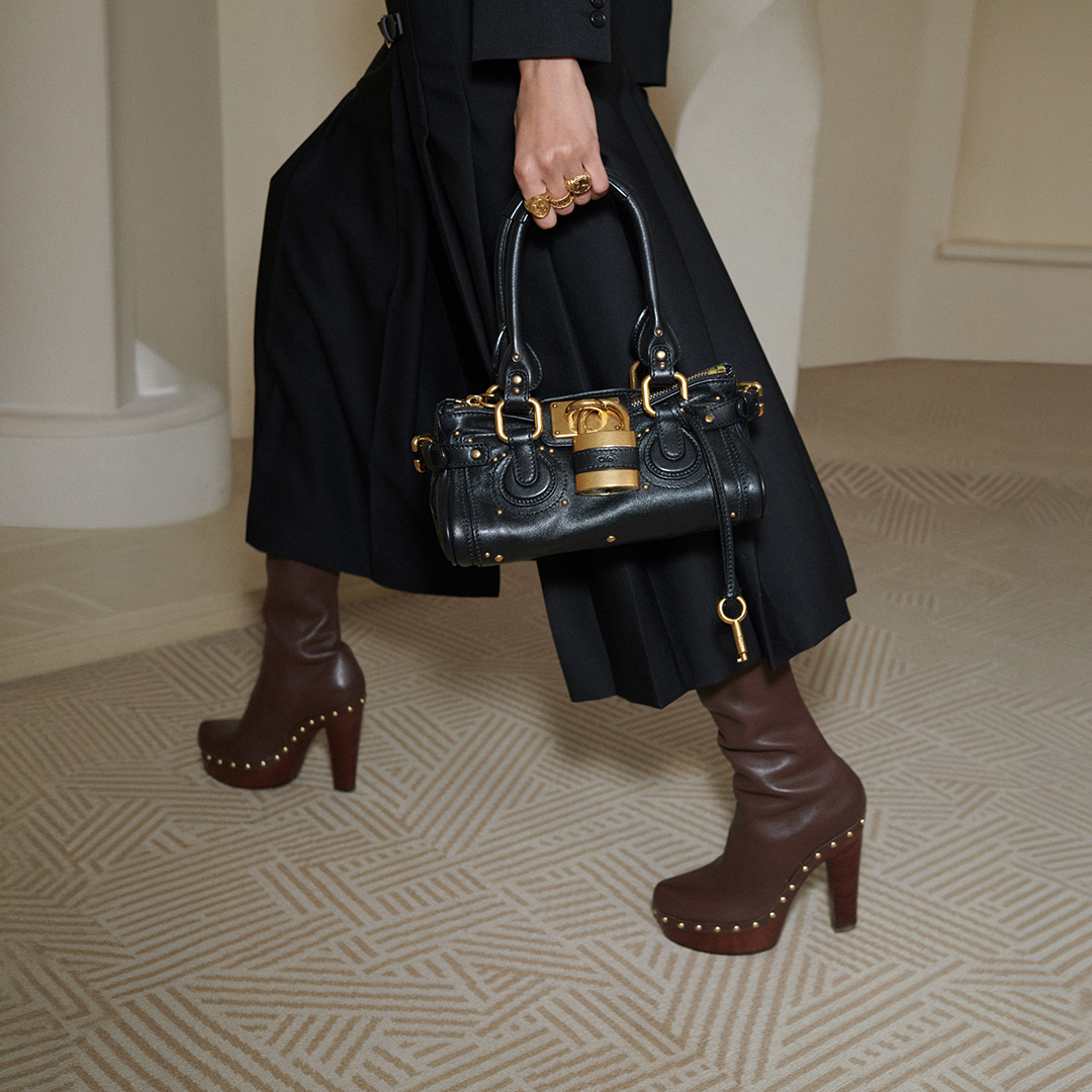 Woman wearing black blazer and pleated skirt carrying black leather handbag with gold hardware and brown studded platform boots