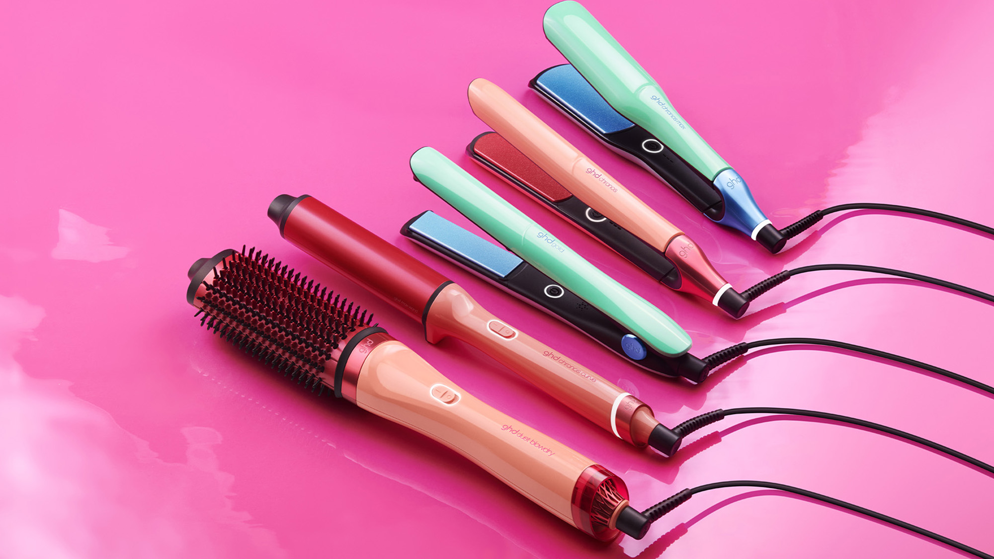 Collection of GHD hair styling tools in pastel colours including straighteners, hair dryer brush and curling irons arranged on pink background