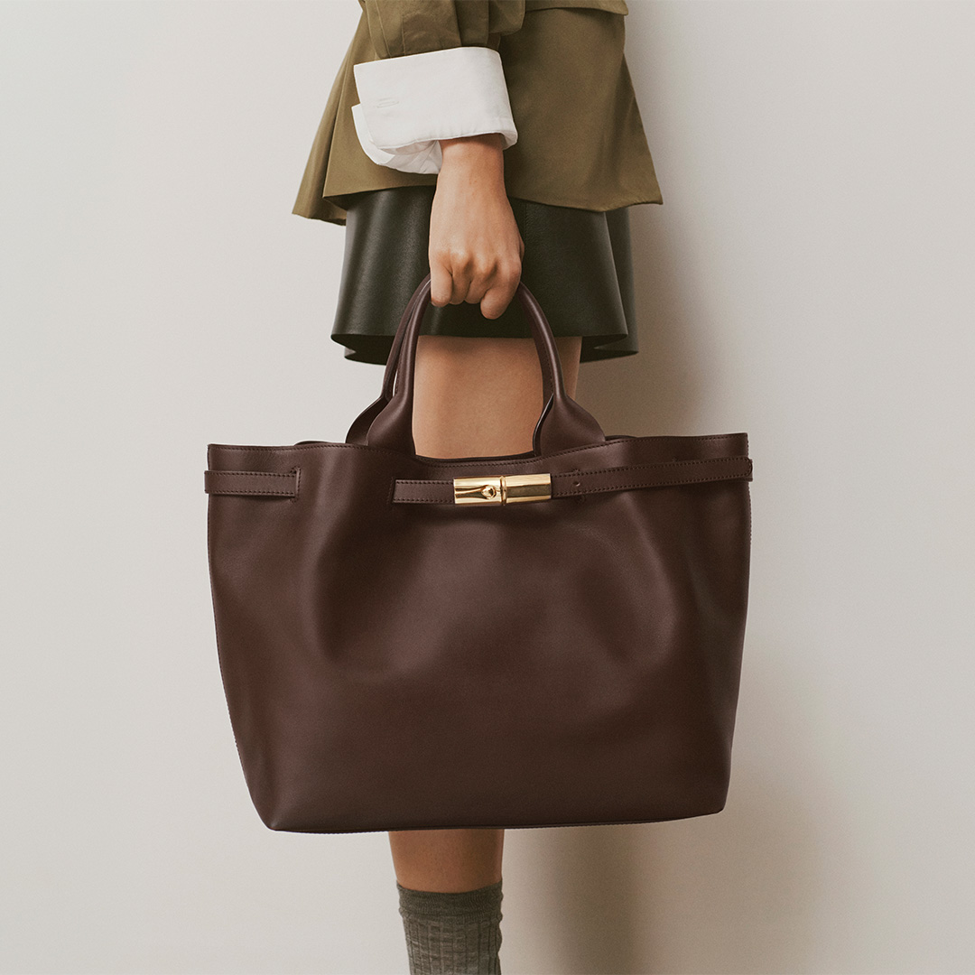 Woman in olive coat holding brown leather tote bag with gold clasp detail and white cuff