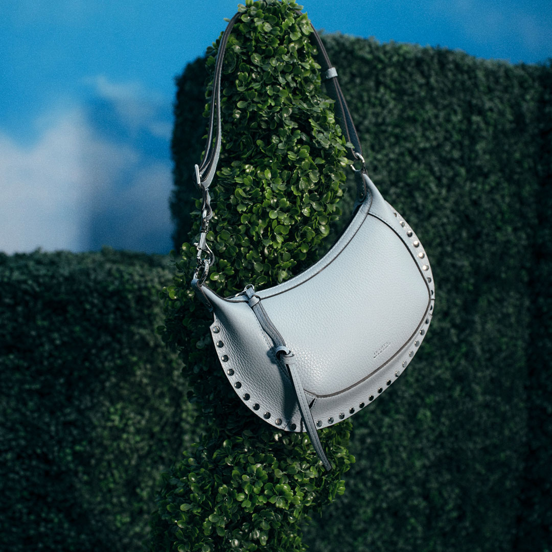 White leather crossbody bag with studded trim and dark navy straps hanging against green foliage