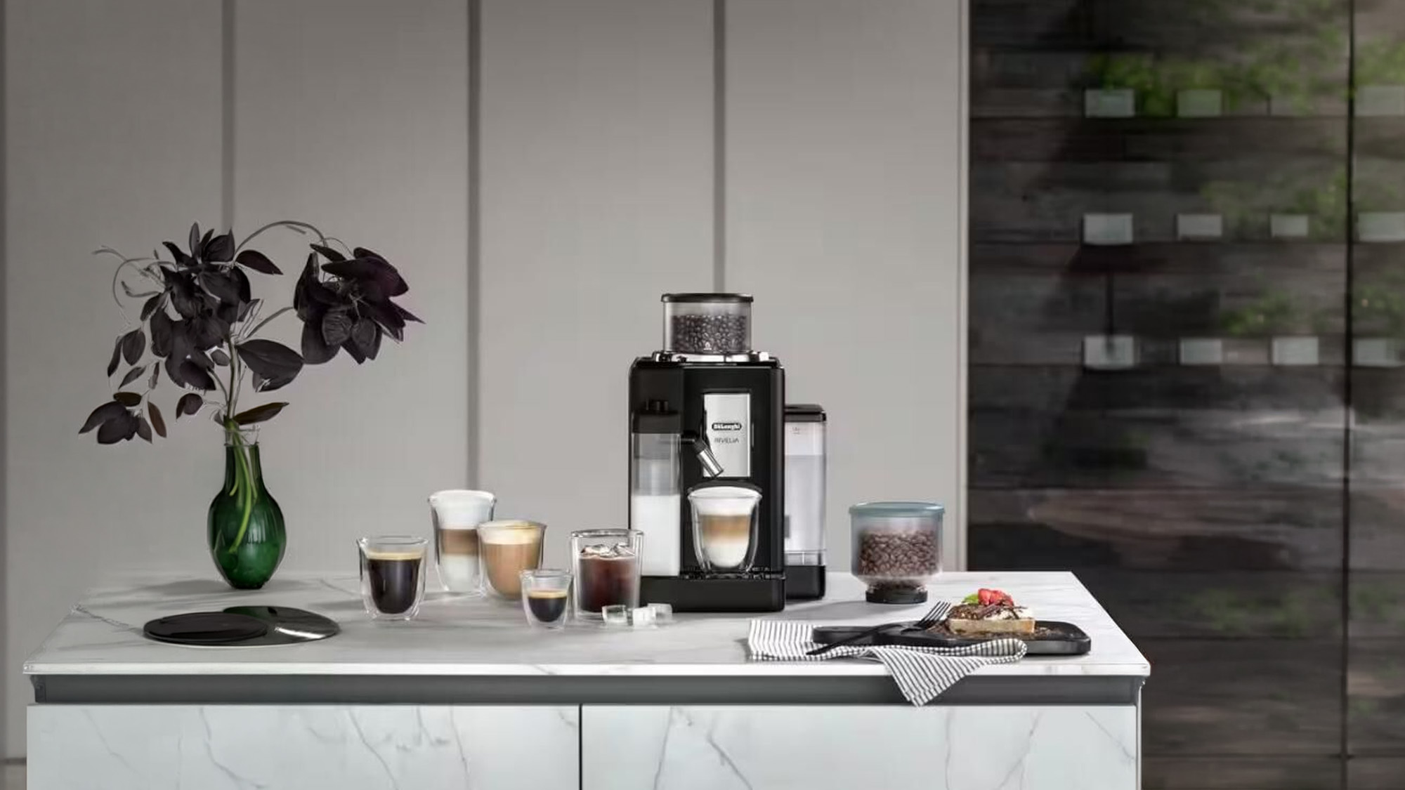 Black Delonghi coffee machine on marble countertop with various coffee cups, coffee beans, dark flowers in green vase, and food items