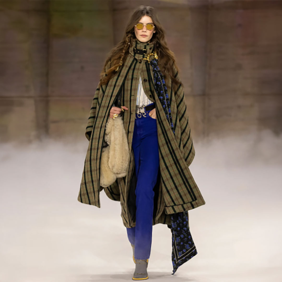 Model walking runway in plaid cape, blue pants, and sunglasses, holding a fuzzy purse.