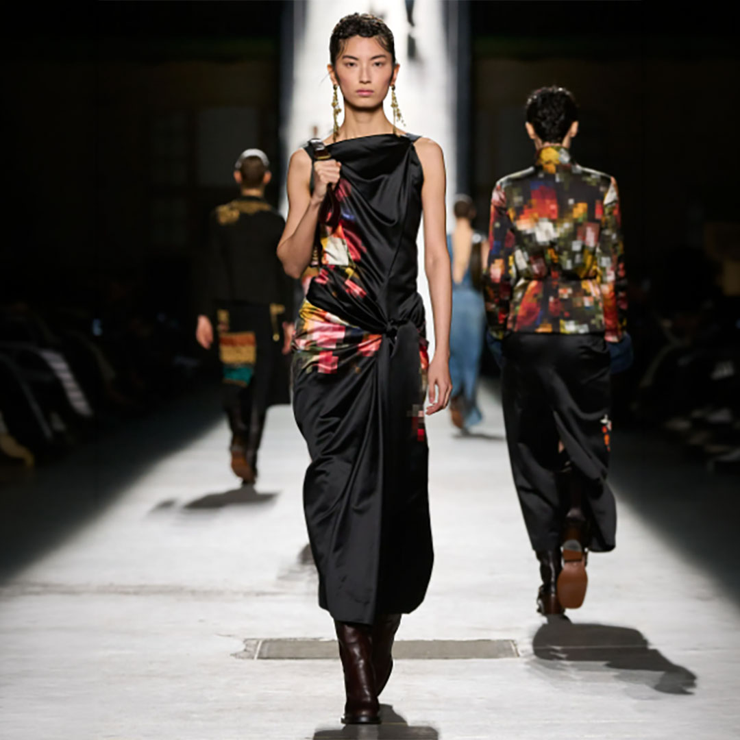 Model walking down a fashion runway wearing a sleeveless black dress with colorful patterns, dark boots, and statement earrings.