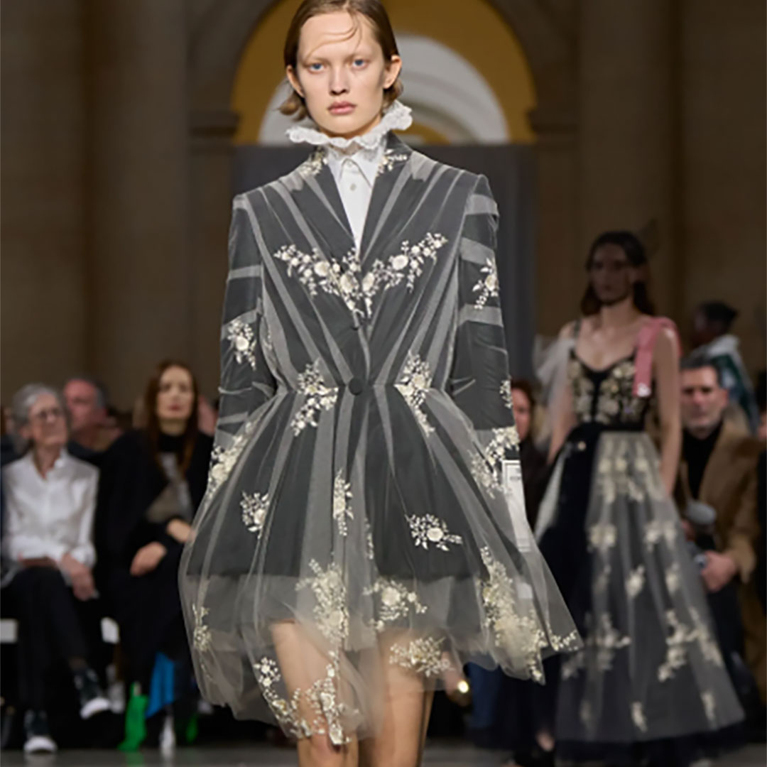 Model walking the runway in a floral-embroidered sheer dress with a structured, professional design. Audience in the background.