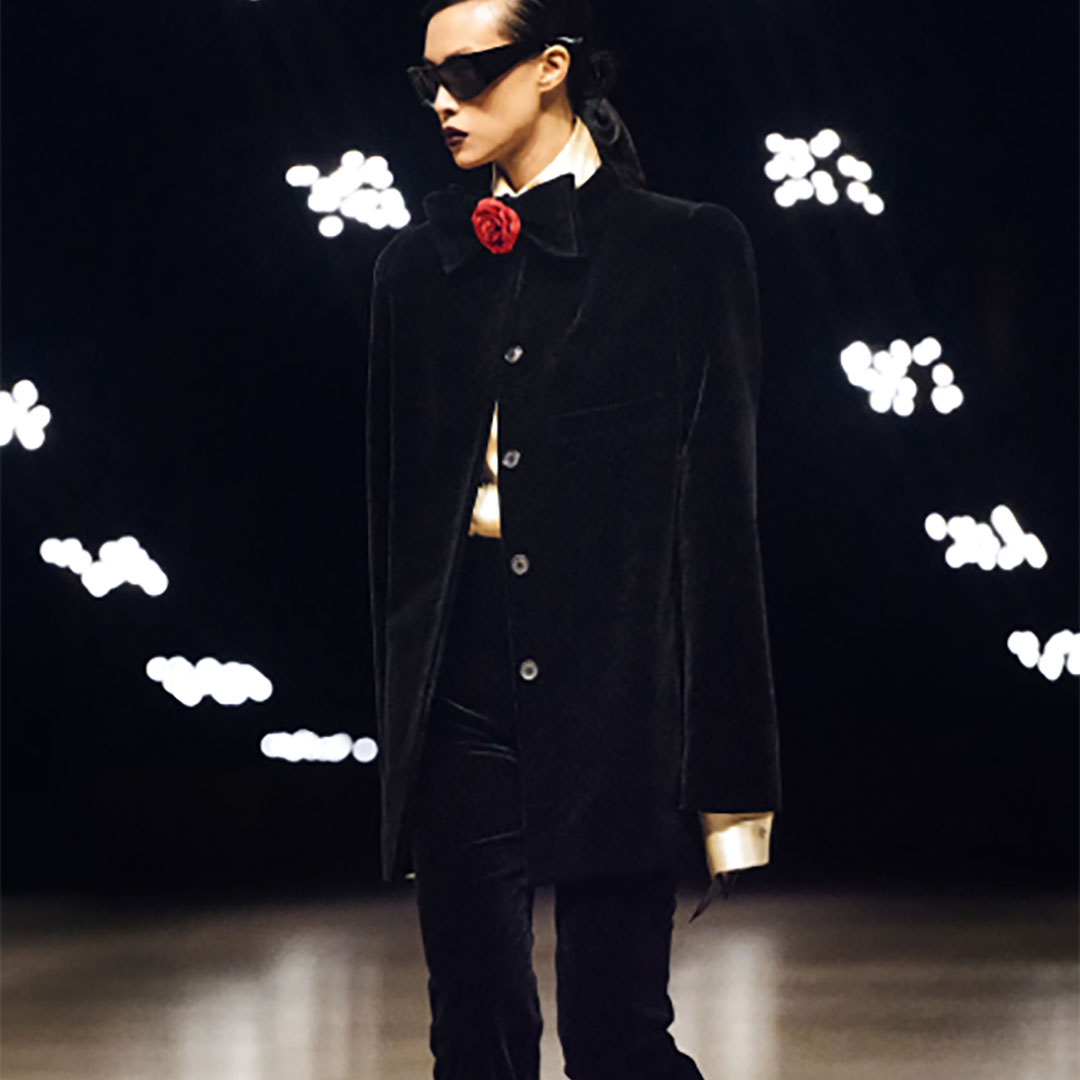 A person in a dark coat with a red rose, wearing sunglasses, walks on a runway with abstract white shapes in the background.