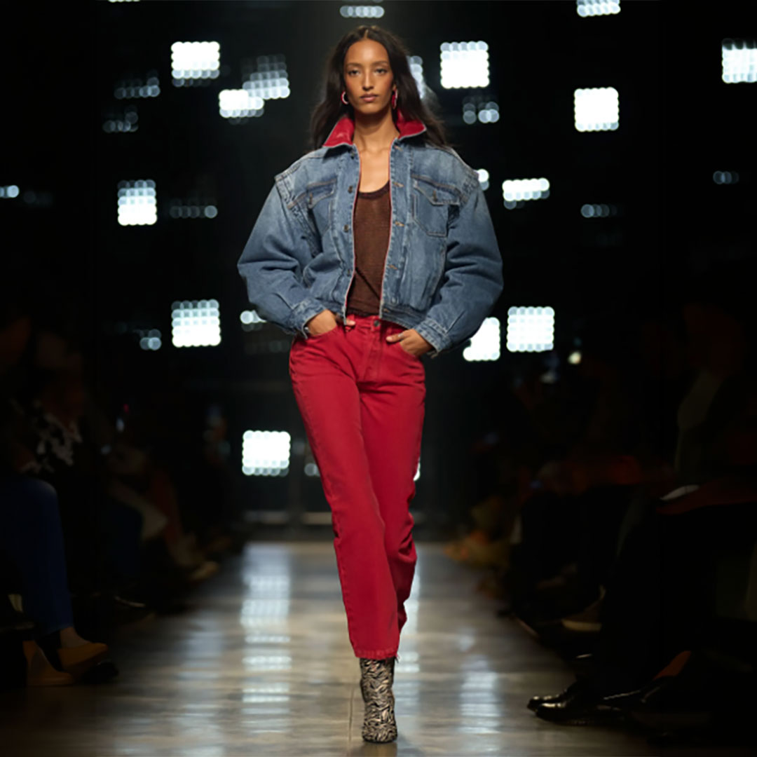 Model walks runway in denim jacket, red pants, and patterned boots against a backdrop of bright lights.