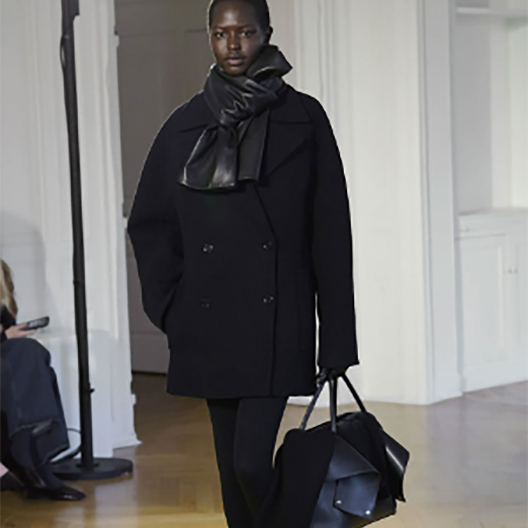 A person models a dark, double-breasted coat with a leather scarf and a large bag, walking indoors.
