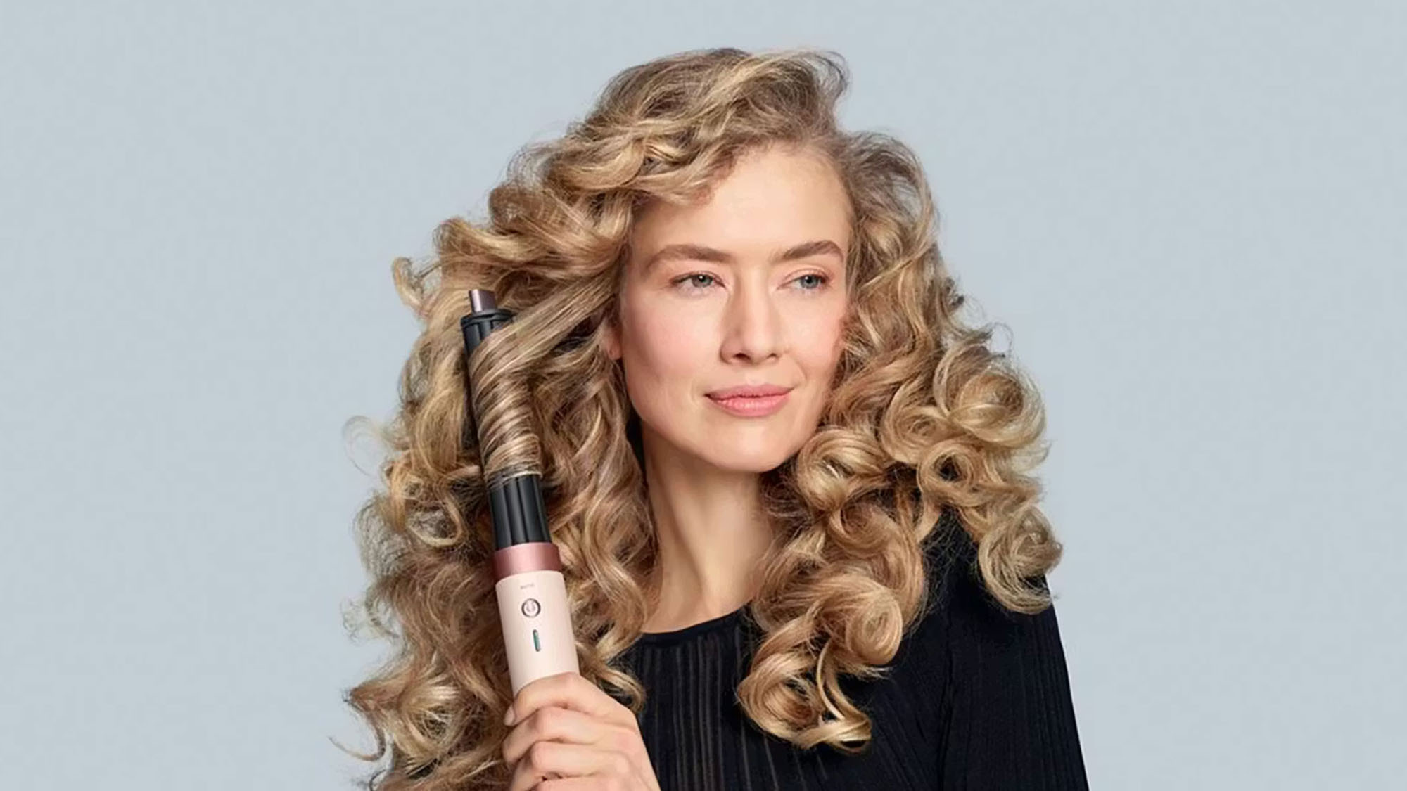 Woman with long wavy blonde hair holding a white and rose gold hair curling tool against her hair