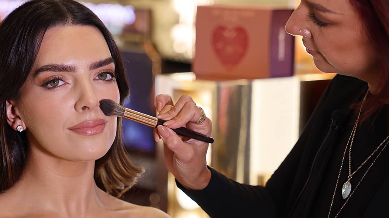 A woman is applying makeup with a brush to another woman's face in a well-lit setting.