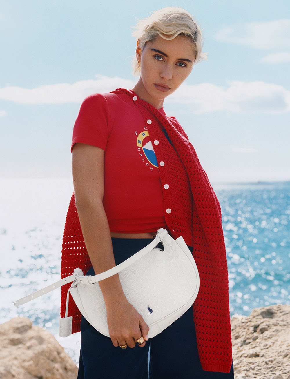 A person in a red outfit stands by the sea, holding a white bag.