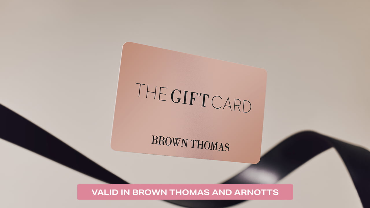 A Brown Thomas gift card is displayed, floating above dark ribbons, with text indicating it's valid in both Brown Thomas and Arnotts.