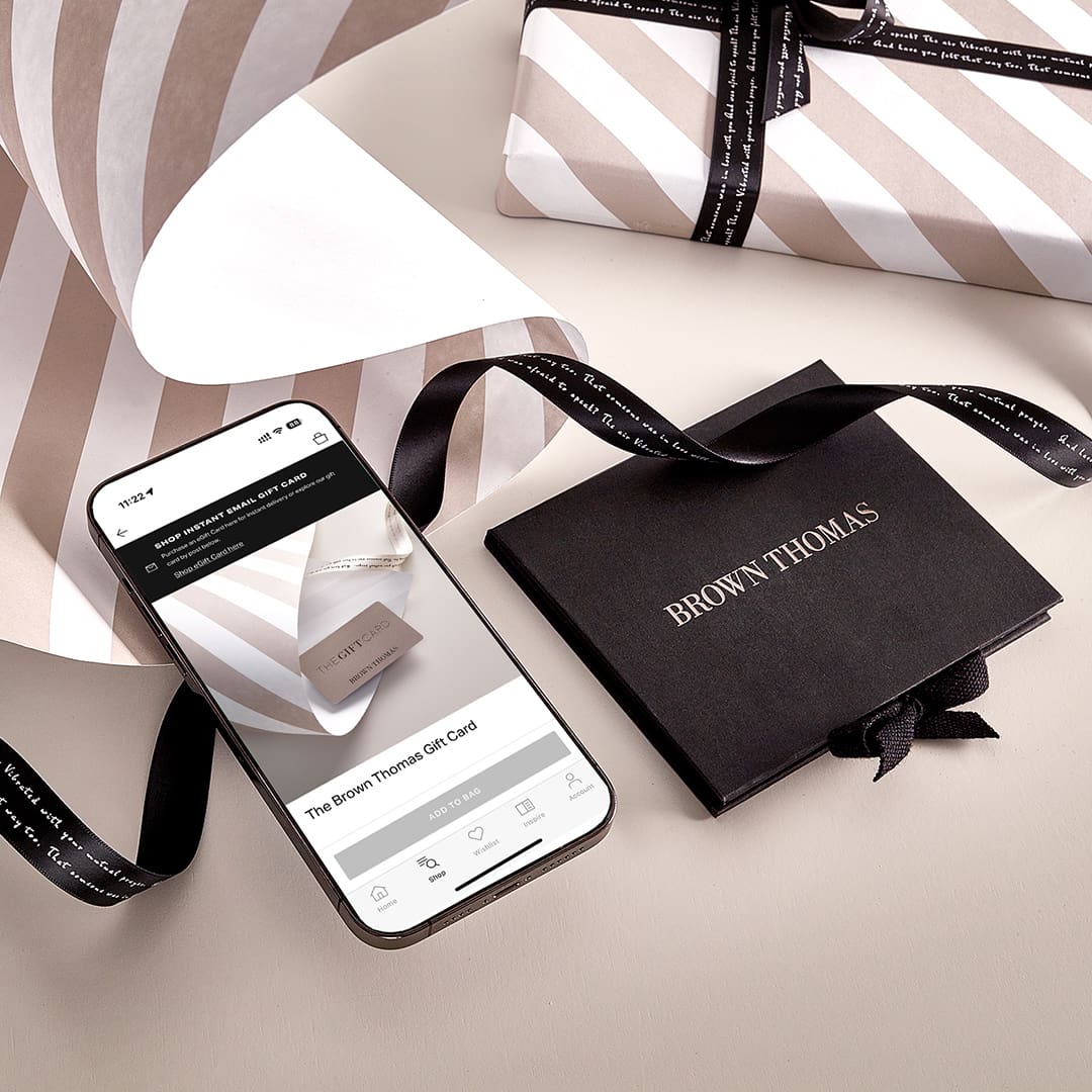 Brown Thomas gift card displayed on smartphone screen next to black branded gift box with black ribbon, on striped background