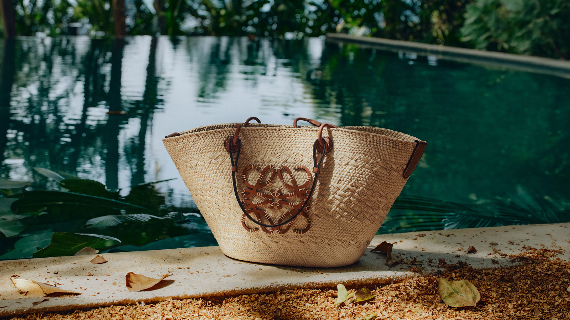Woven straw tote bag with brown leather handles and embroidered detail on sandy ground by water