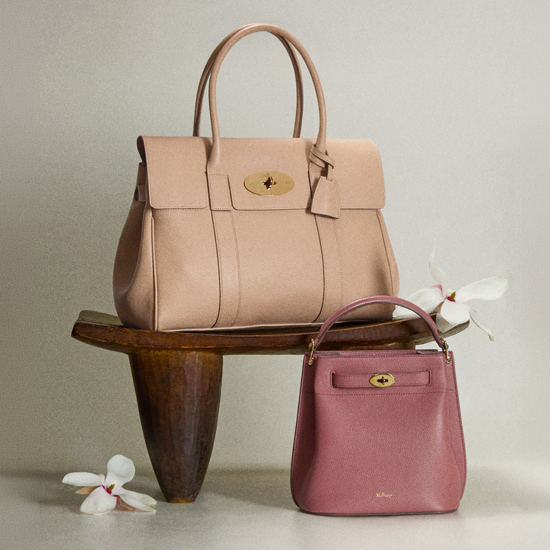Two leather handbags displayed on wooden bench: beige tote with gold clasp and pink crossbody bag, with white flowers nearby