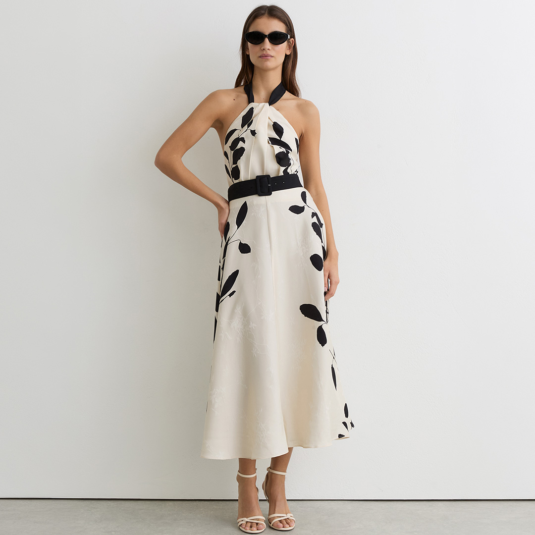 Woman wearing cream halterneck dress with black leaf print and black belt, styled with sunglasses