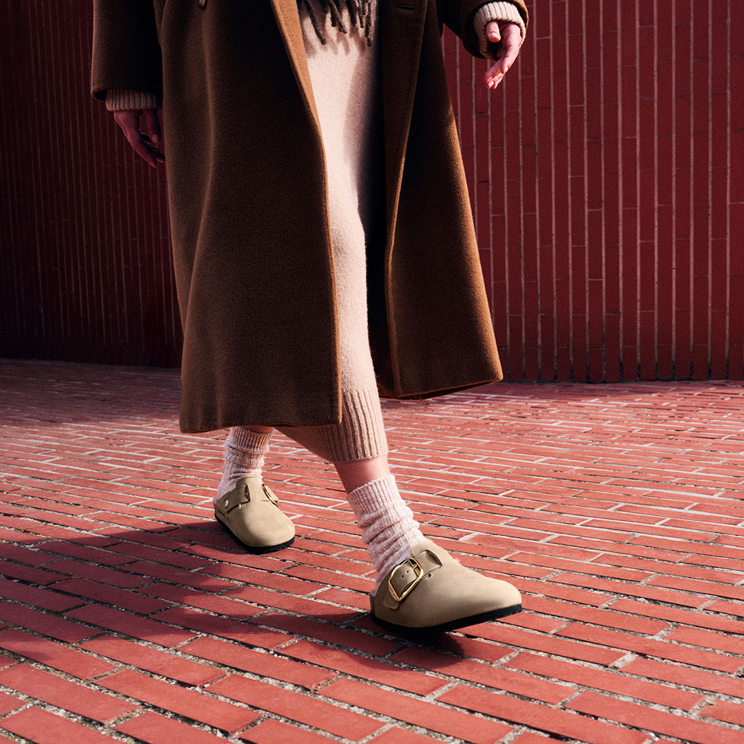 Person wearing cream Birkenstock sandals with white socks and brown camel coat standing on red brick pavement