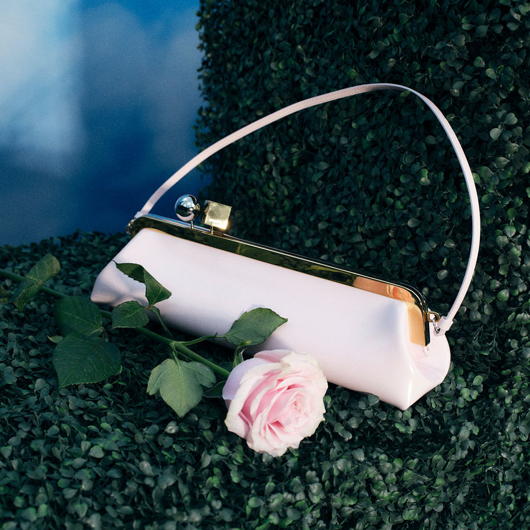 White structured handbag with black trim and pale pink handle, displayed with pink rose and green foliage against blue backdrop.
