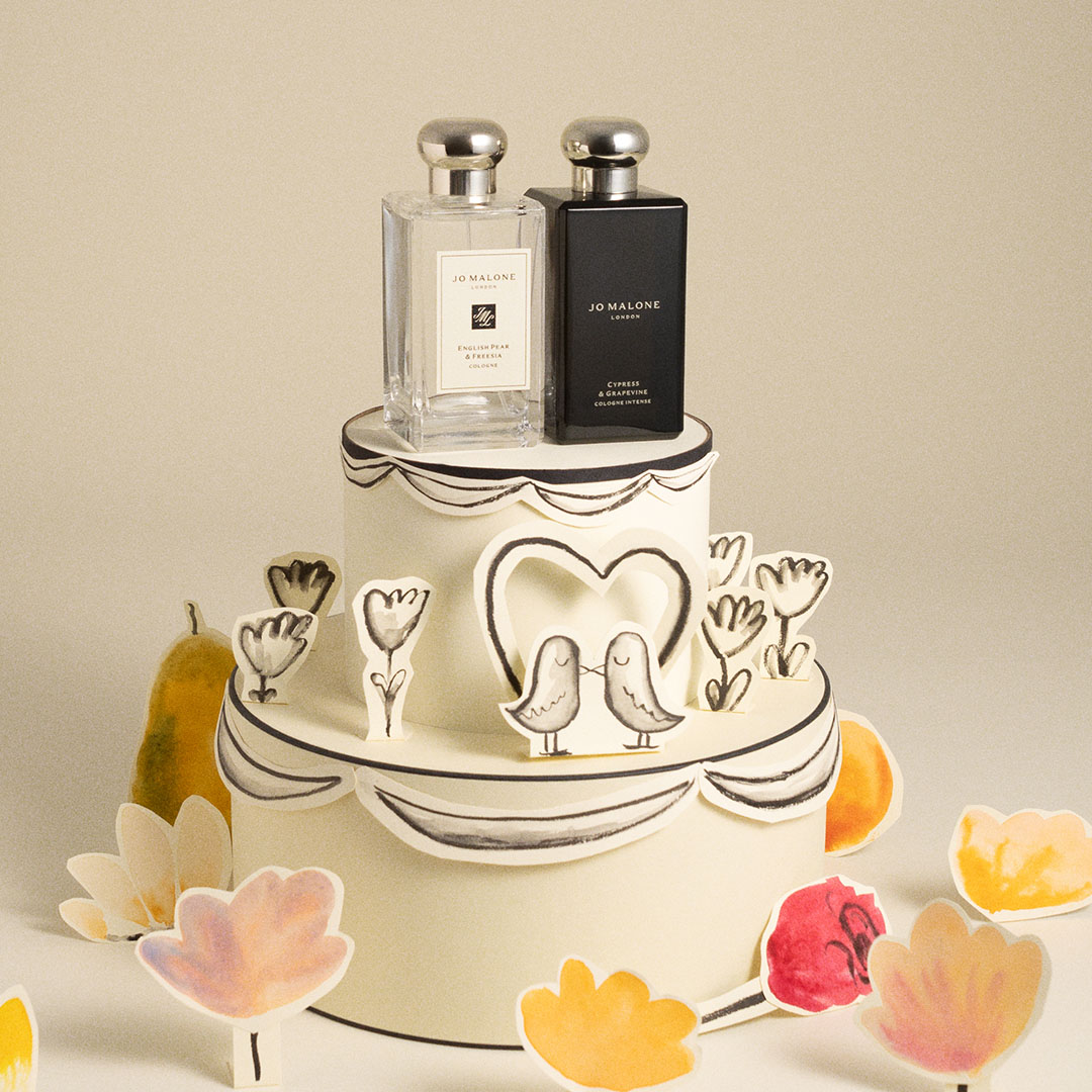 Decorative tiered display cake with illustrated hearts and birds, topped with Jo Malone fragrance bottles in clear and black packaging.