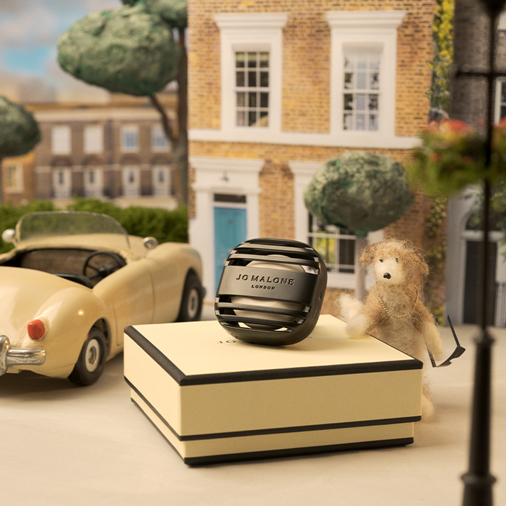 A Jo Malone perfume sits on a box beside a teddy bear, with a vintage car and brick building in the background.