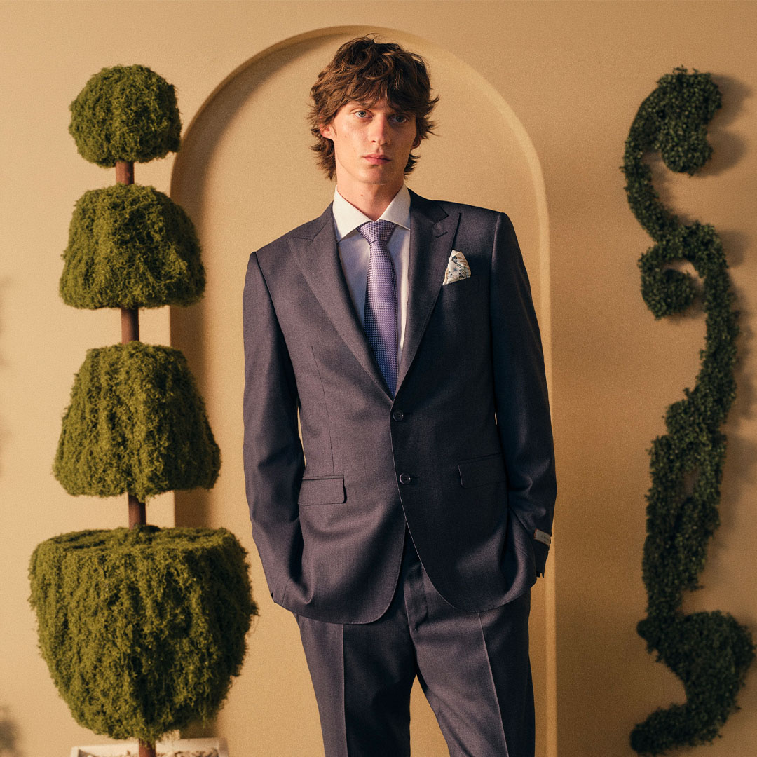 Man wearing navy suit with purple tie and pocket square, standing against beige wall with topiary trees.