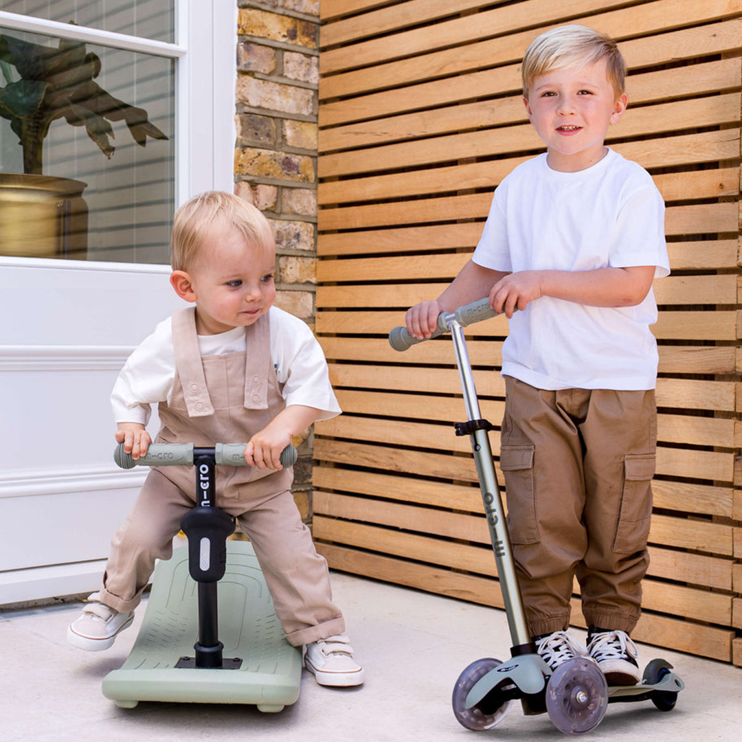 Two young children riding Micro Scooters outside a brick house with wooden gate