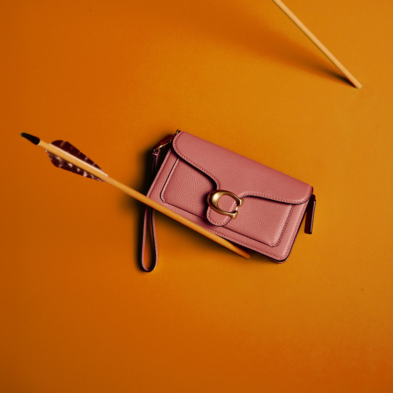 A pink wristlet with a gold buckle is placed against a red background, with an arrow lying across it.