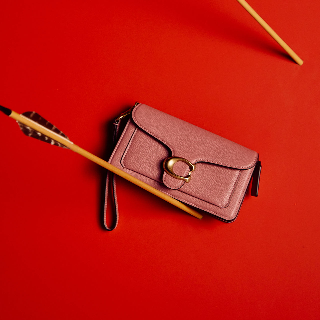 A pink wristlet with a gold buckle is placed against a red background, with an arrow lying across it.