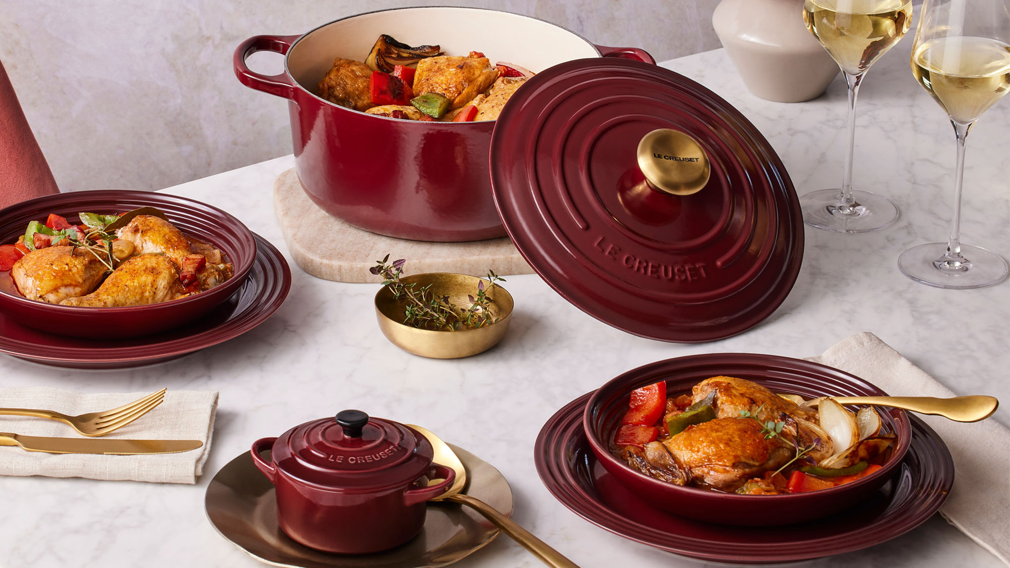 A table set with red cookware featuring roasted chicken, gold utensils, two glasses of white wine, and a small red pot.