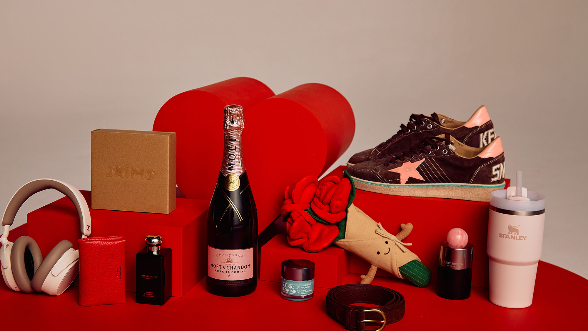 A collection of gifts including headphones, a bottle of champagne, sneakers, cosmetics, a plush rose, and a branded tumbler on a red surface.