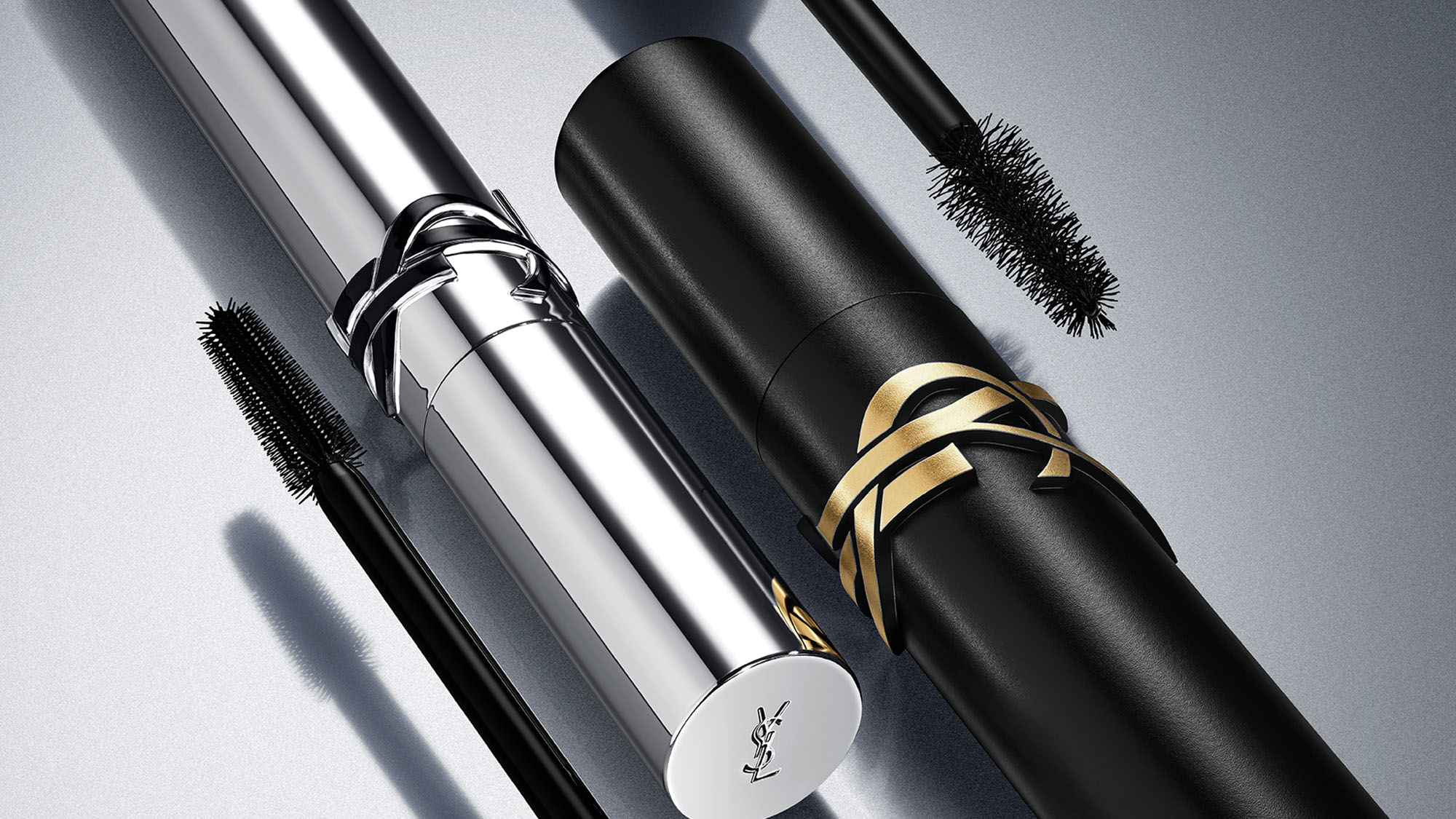 Two mascara tubes, one silver and one black with gold accents, placed on a reflective surface with their brushes beside them.
