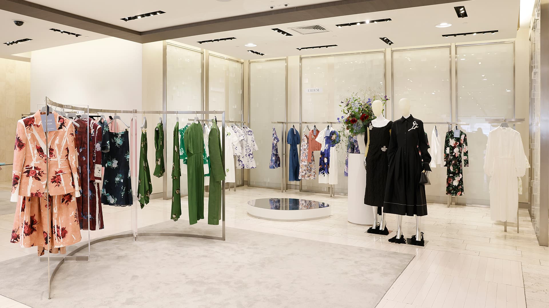 A stylish clothing boutique featuring modern displays of women's dresses and outfits. Garments in floral prints, solid greens, and black hang on racks. A small platform is centrally placed, with mannequins on the right displaying black dresses. The store's decor is minimal, with light-coloured walls and flooring.