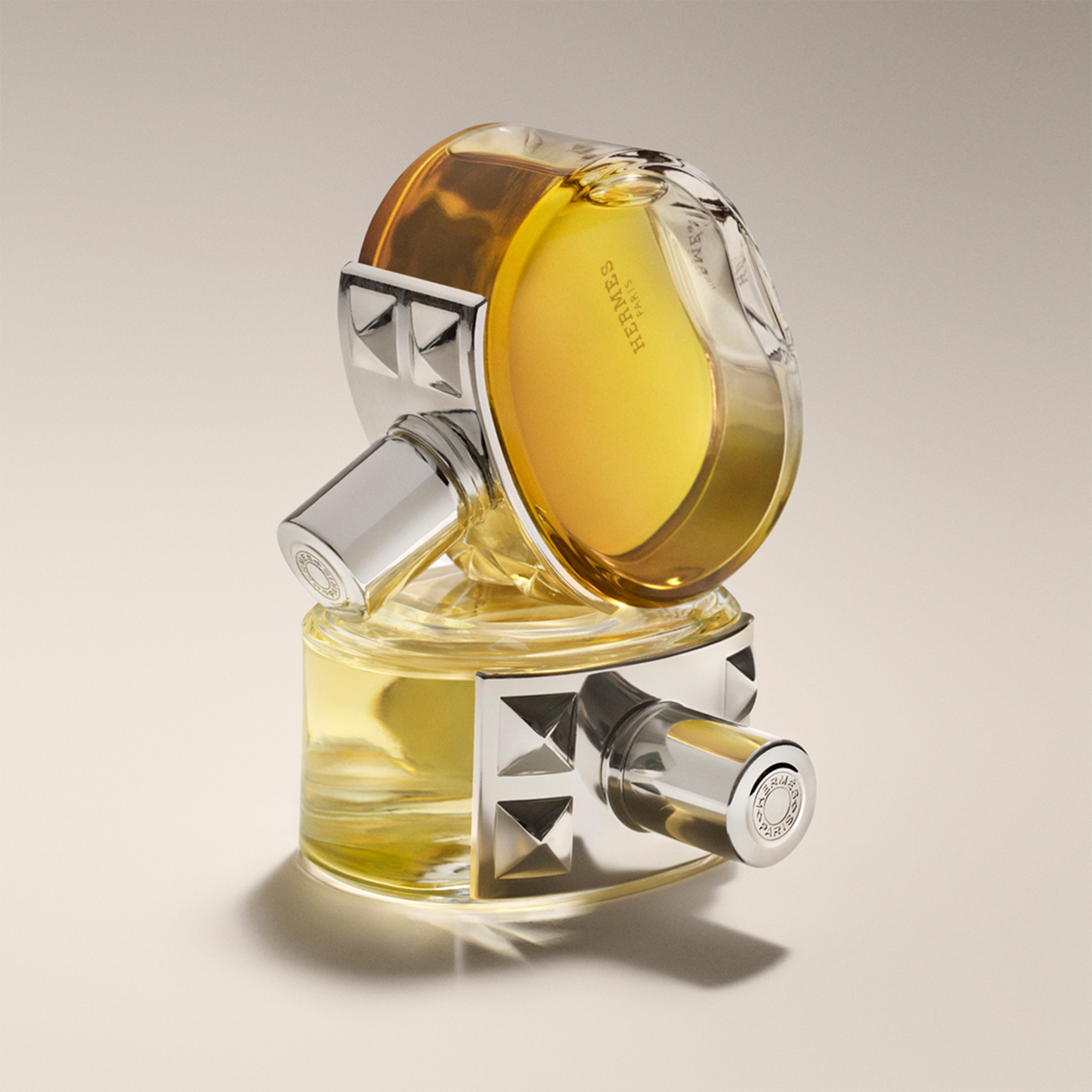 A round, dual-chamber perfume bottle with silver accents and amber liquid, creatively designed and positioned on a beige background.
