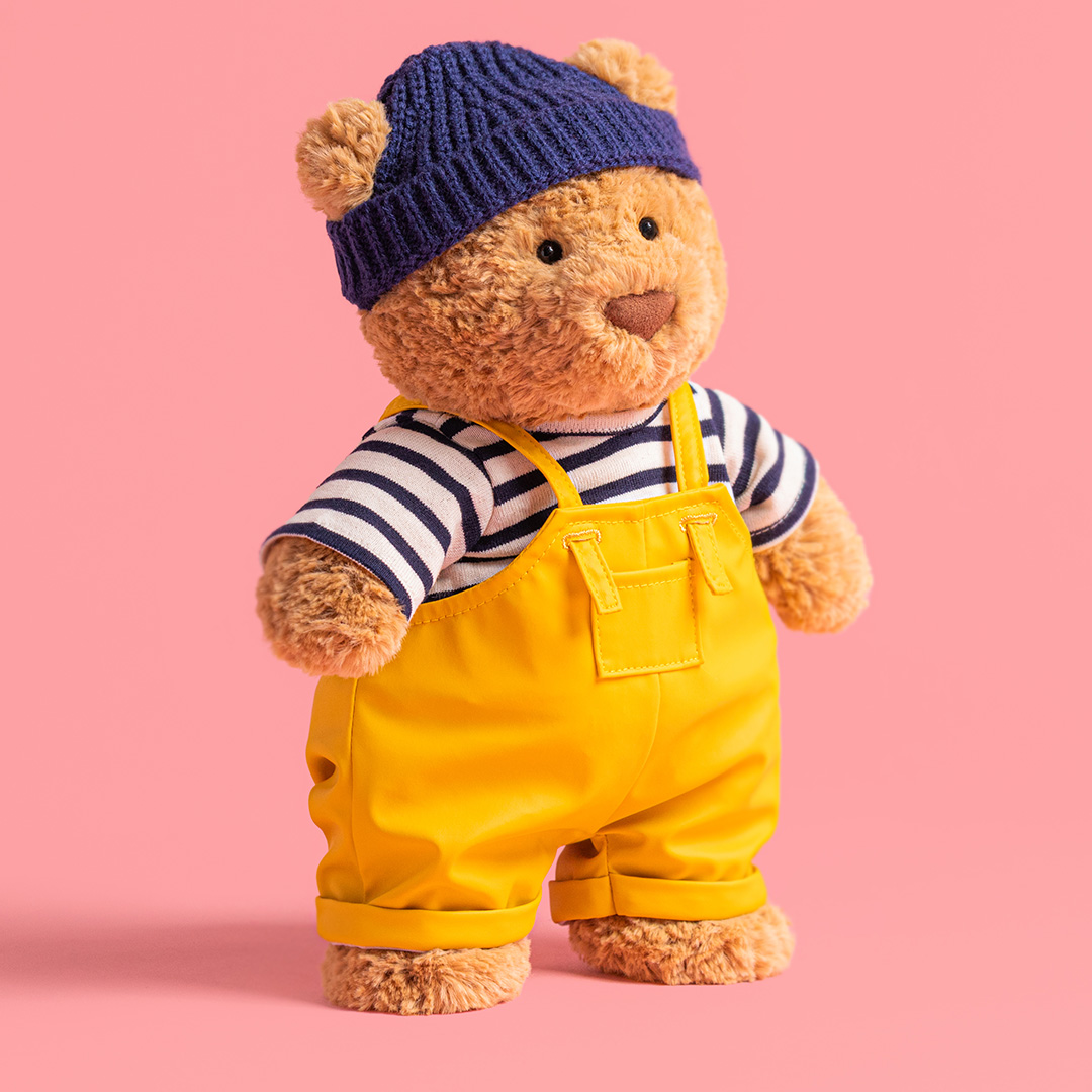 Tan teddy bear wearing navy knit hat with ears, navy and white striped shirt, and yellow dungarees.