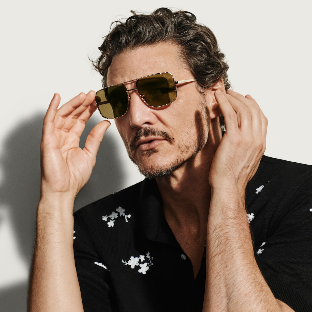 A man with short hair, wearing sunglasses, adjusts them confidently. He is dressed in a dark shirt with light-coloured patterns. The background is plain, focusing attention on his thoughtful expression and stylish appearance.