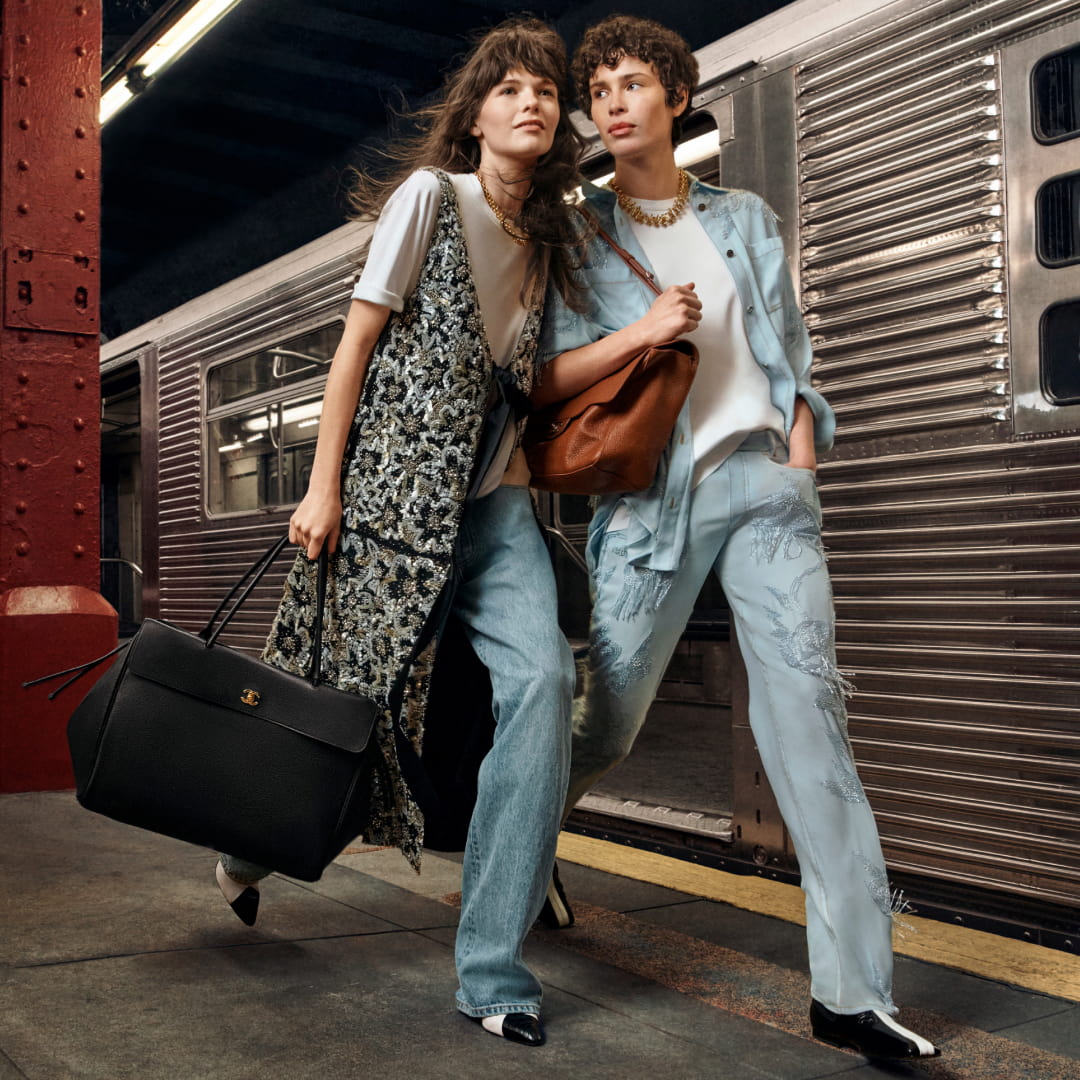 Two people stand on a subway platform. One wears a patterned long vest over a white shirt and jeans, holding a black bag. The other wears a light blue suit with a white top, holding a brown bag. A subway train is visible in the background.