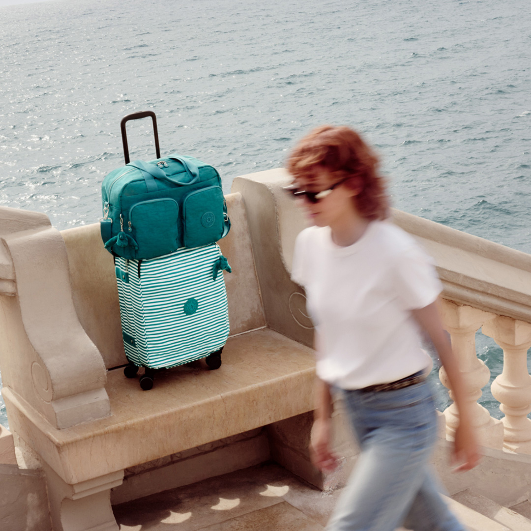 Woman running towards teal and striped carry-on luggage on seaside balcony overlooking calm ocean waters