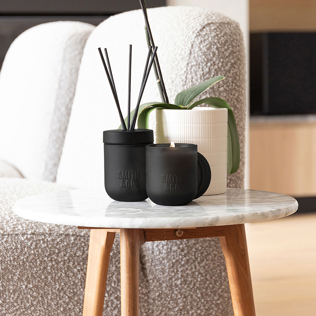 Smith Co black reed diffusers and cream candle arranged on marble table beside grey upholstered chair