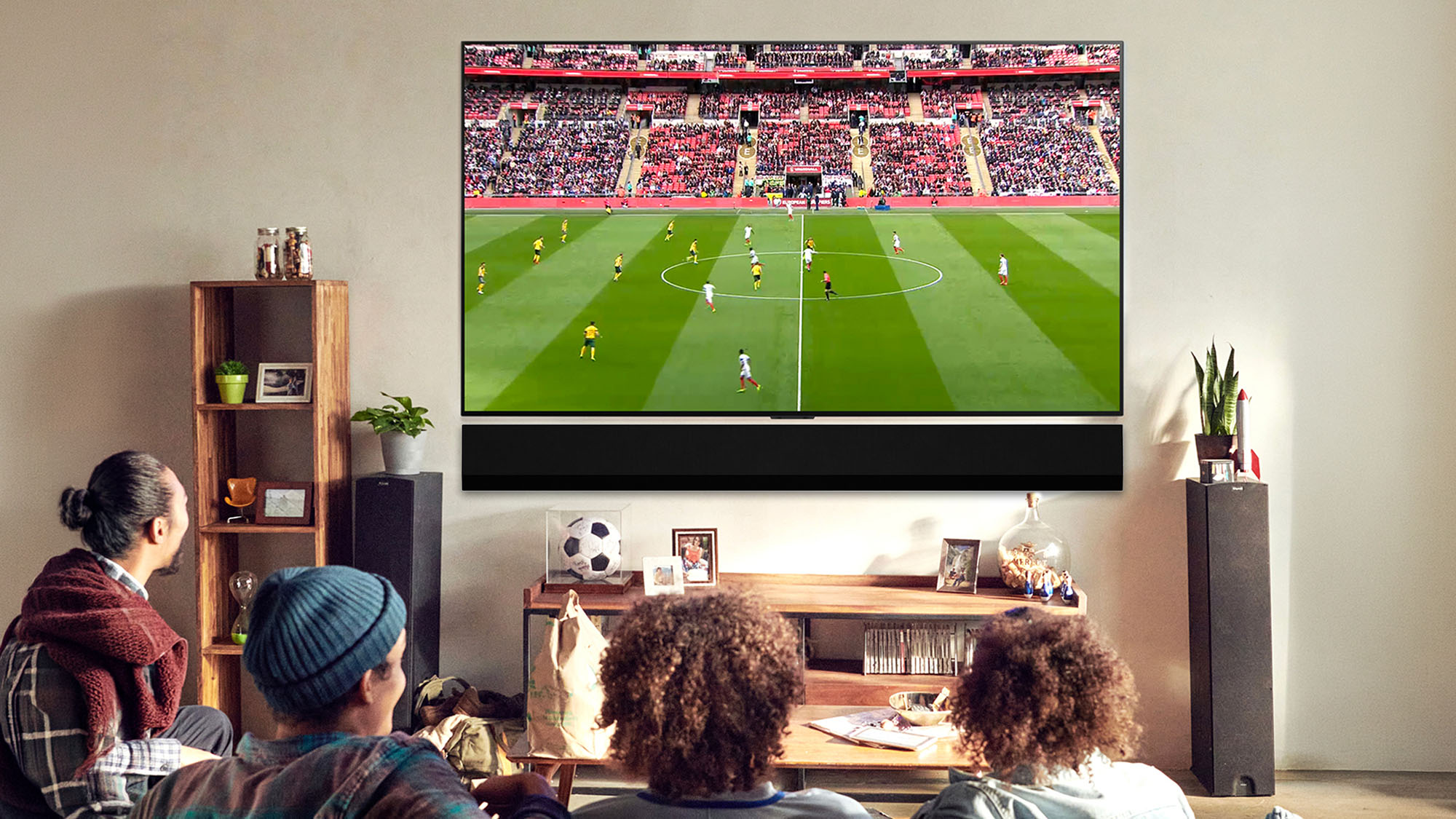 Group of people watching football match on large wall-mounted television in living room