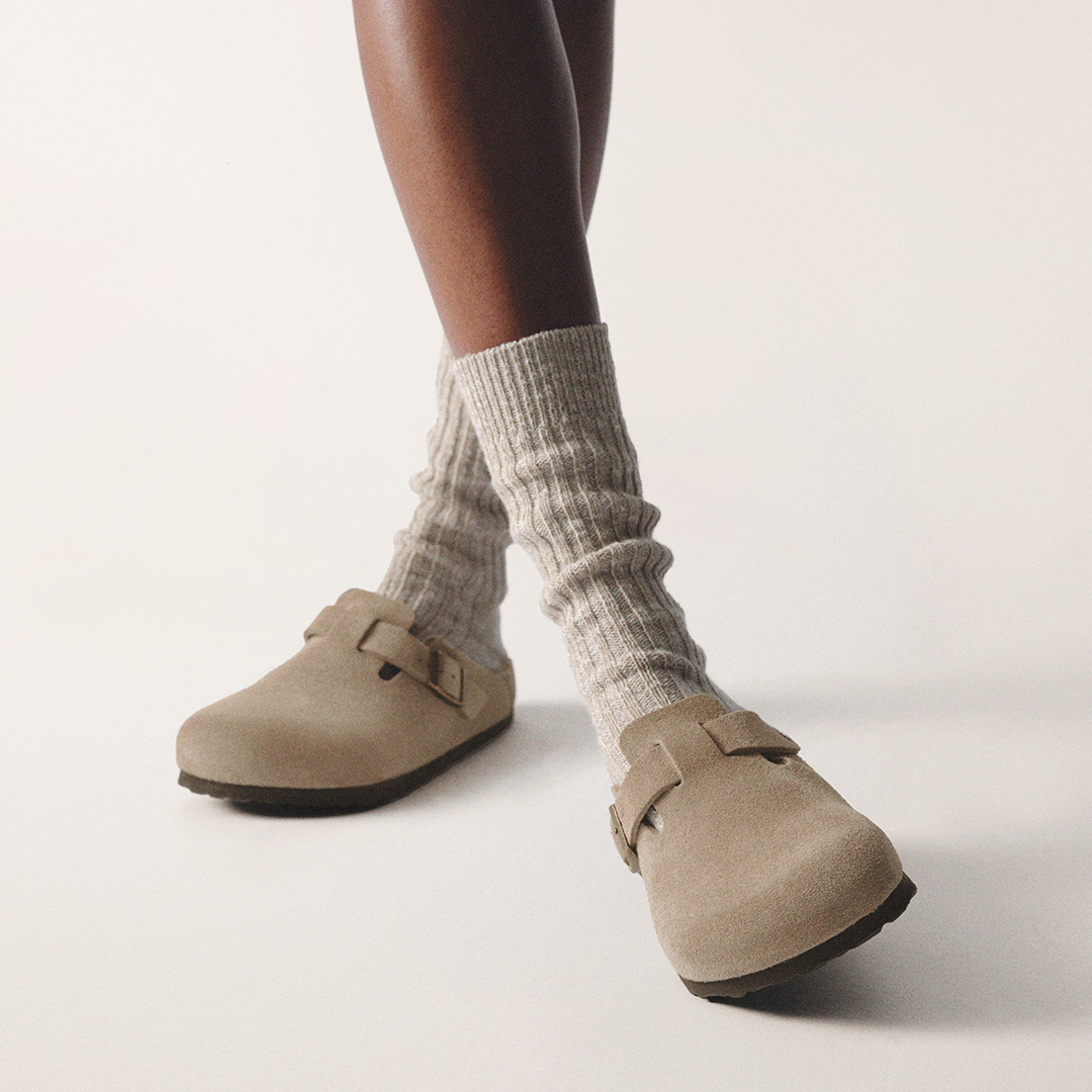 Person wearing cream ribbed socks and beige suede slip-on clogs with buckle detail on white background