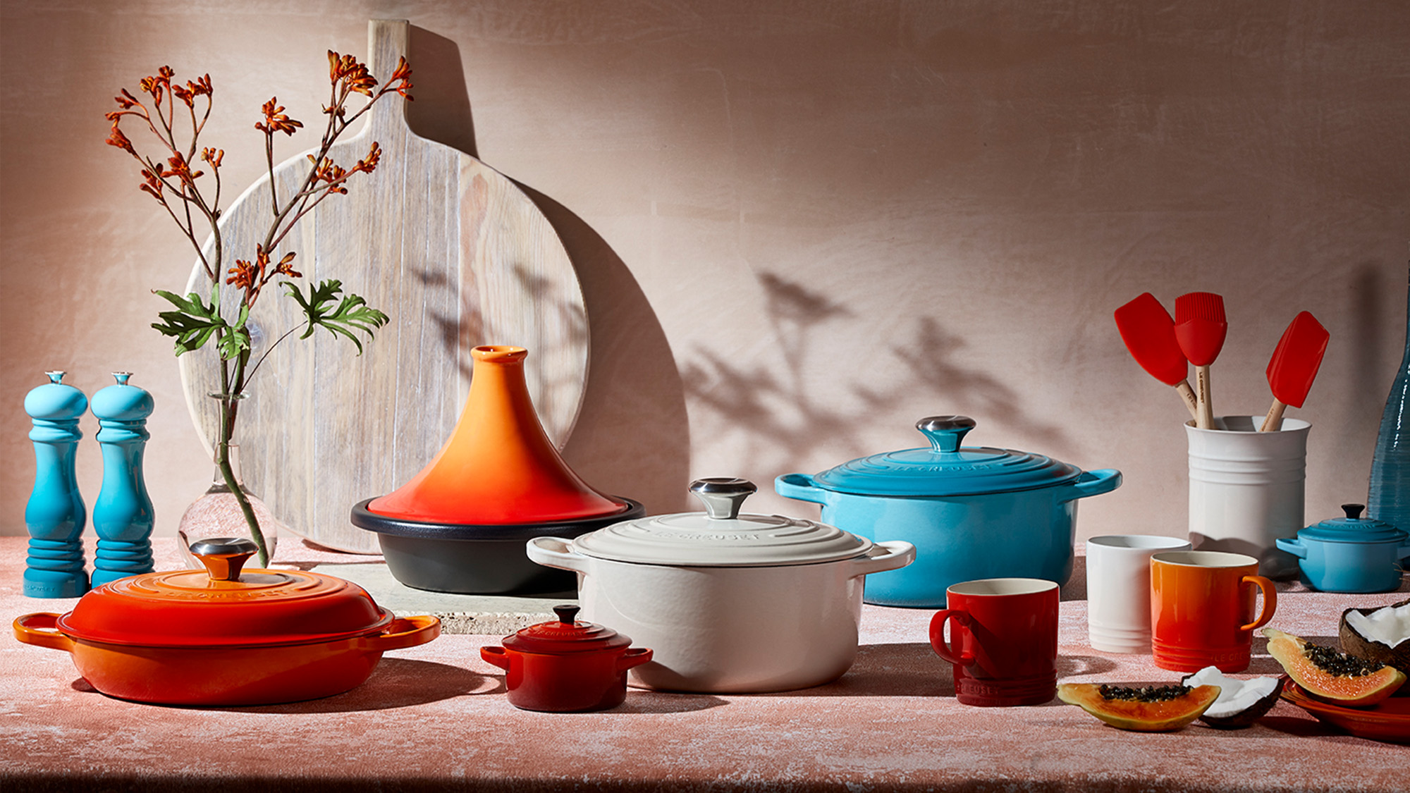 Colourful home cookware and kitchen accessories including orange and blue pots, white casserole dishes, salt and pepper mills, and red utensils arranged on neutral surface