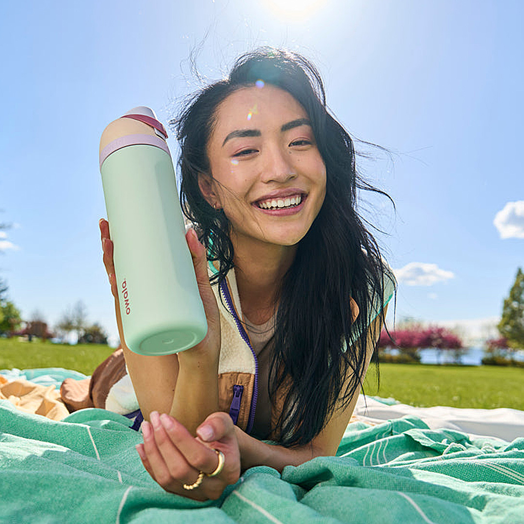Woman holding mint green water bottle with burgundy trim on picnic blanket outdoors, smiling in sunny park setting